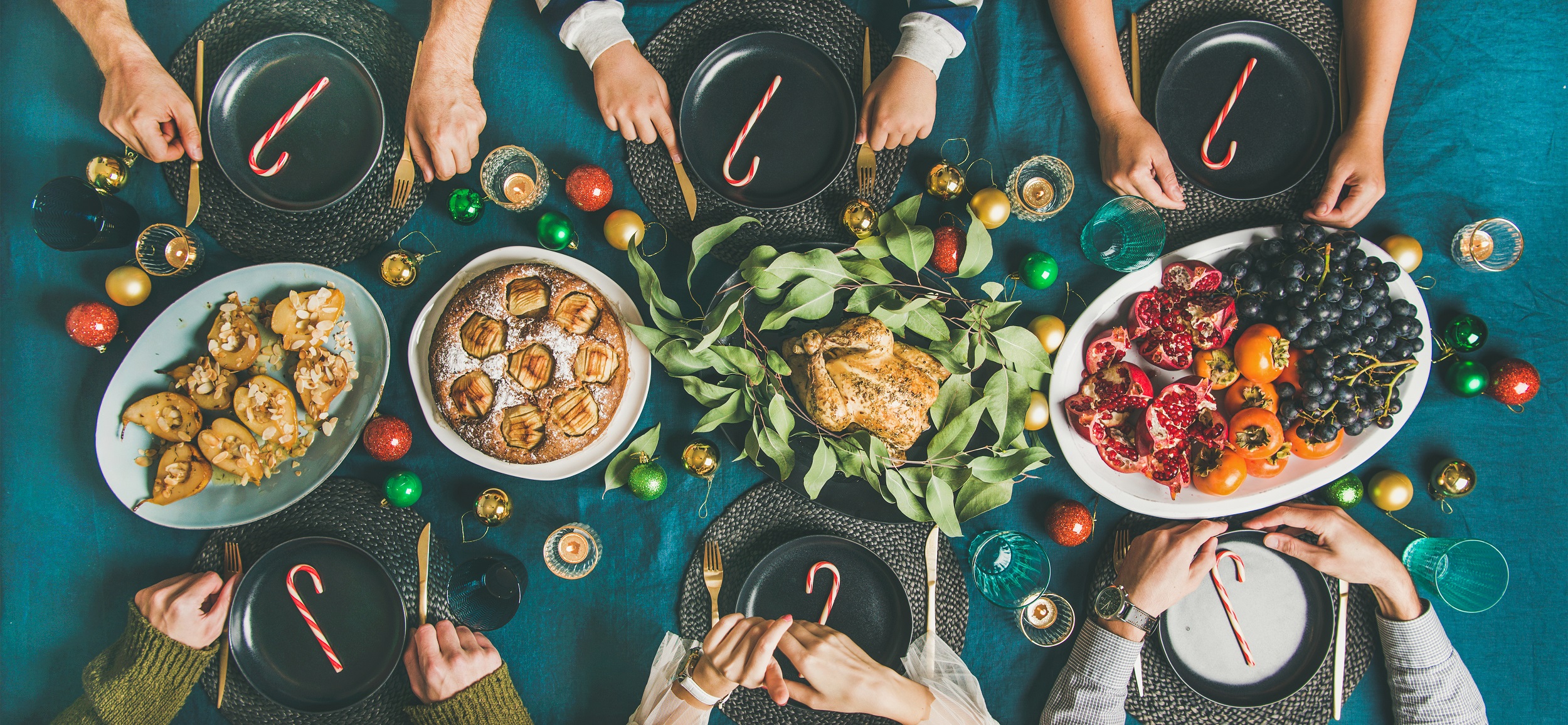 Company of friends gathering for Christmas or New Year party dinner at festive table. Flat-lay of people sitting with plates and ready to start celebrating holiday together, top view, wide composition